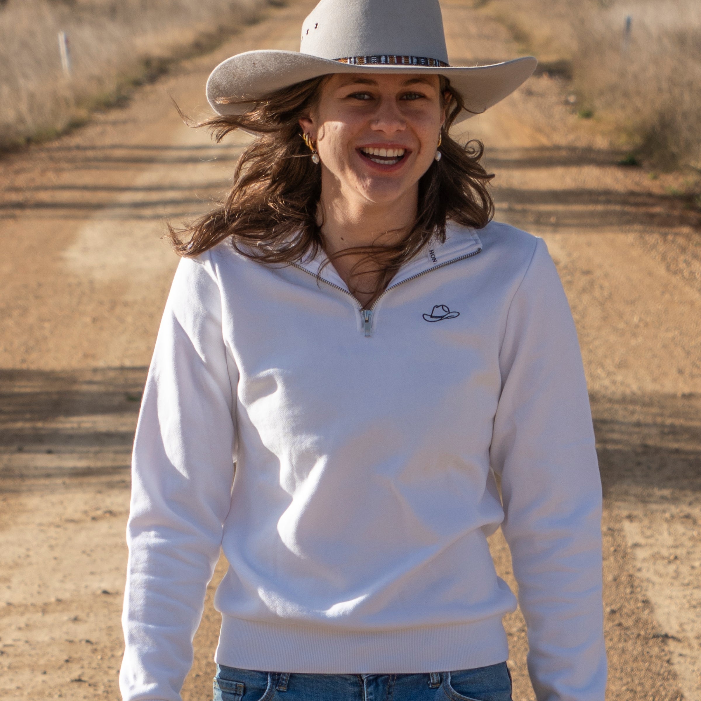 Woman wearing a white sweatshirt and cowboy hat on a dirt road with trees in the background