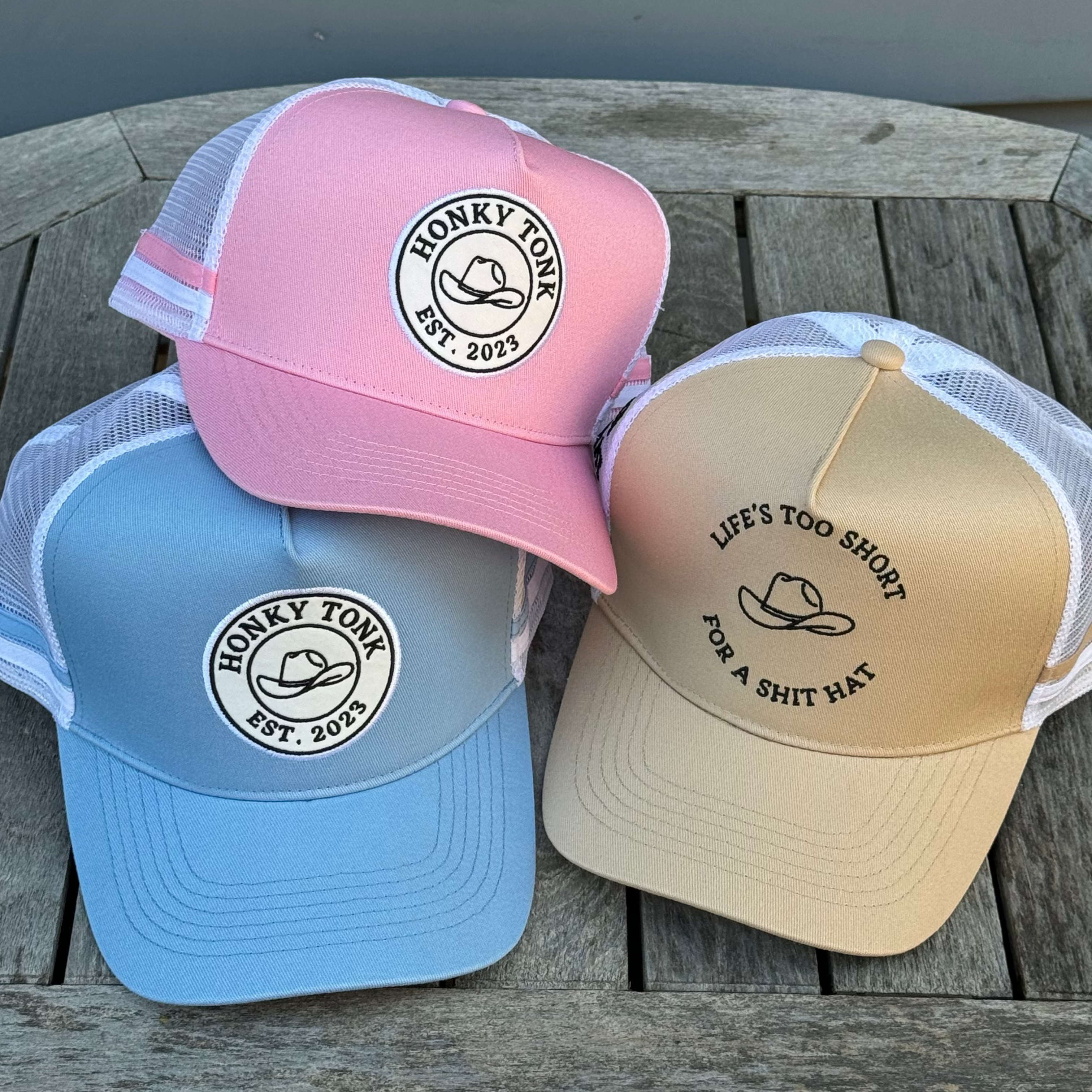 Three caps in pink, blue, and beige on a wooden surface with text and logos.