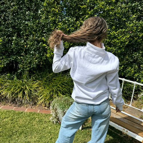 Person in a white jacket and blue jeans standing outdoors with greenery in the background